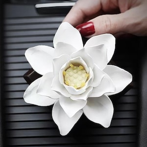 May include: White ceramic car air freshener shaped like a lotus flower with a yellow center. The flower is sitting on a black surface.