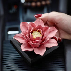 May include: A pink ceramic lotus flower air freshener with a gold center, sitting on a black square base. The air freshener is placed in a car.