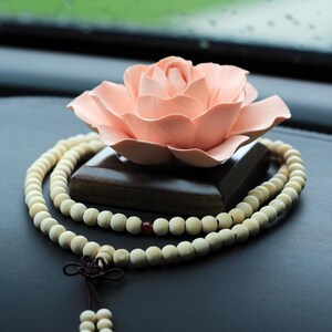 May include: A light pink lotus flower sits atop a dark wooden block, with a cream-colored beaded necklace draped around it. The necklace has a dark brown tassel and a single red bead. The background is dark.