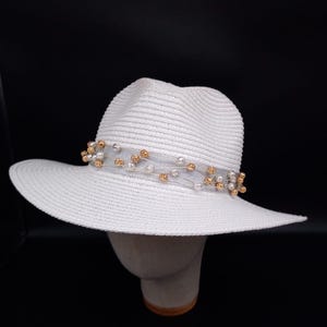 May include: A white fedora hat with a wide brim and a textured woven surface. The hat features a decorative band made of sheer mesh fabric adorned with small, scattered metallic gold and silver spherical beads encircling the crown.