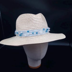 May include: A beige straw fedora hat featuring a light blue sheer ribbon band wrapped around the crown. The ribbon is embellished with small, translucent blue and clear beads woven into the fabric, creating a textured, decorative accent.