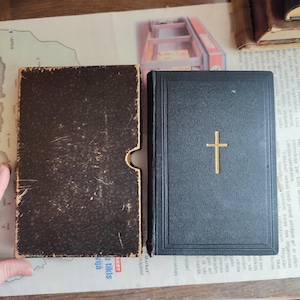 May include: A black book with a gold cross emblem and a matching case. The book is closed and has a textured cover. The case is open and has a worn, dark brown appearance. The items are on a map.