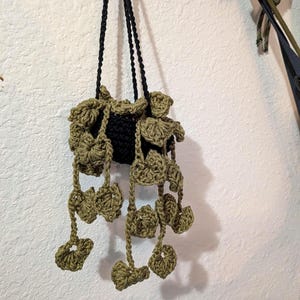 May include: A handmade crochet plant hanger in black and olive green. The plant holder is suspended by two black cords and features a decorative, leafy design around the top and hanging down. The crochet work is detailed and textured, creating a unique, bohemian aesthetic.