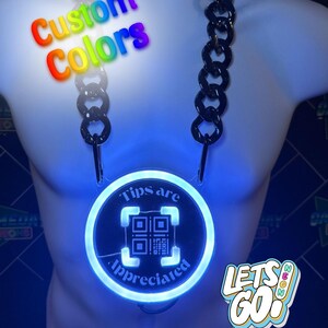 Può includere: Una collana a catena nera con un cartello LED rotondo blu chiaro che dice "Tips are appreciated" con un codice QR al centro. Il cartello è illuminato e la catena è appesa a un manichino bianco.