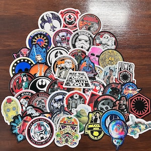 Może przedstawiać: Zbiór kolorowych naklejek z motywem Gwiezdnych Wojen przedstawiających różne postacie, statki kosmiczne i logo. Naklejki są okrągłe i mają błyszczące wykończenie.