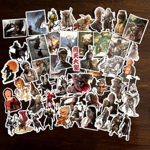 Puede incluir: Una colección de pegatinas coloridas con varios personajes y diseños, incluyendo un personaje rojo y blanco con el texto "魔天大聖" en rojo. Las pegatinas están sobre una superficie de madera marrón.