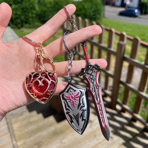 Peut inclure: Trois porte-clés : un charm en forme de cœur doré avec une gemme rouge, un bouclier argenté avec un emblème rose et une épée avec des accents rouges. Les porte-clés sont suspendus à des anneaux argentés et dorés.