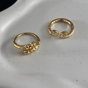 May include: Two gold rings with intricate designs. The rings are on a white surface.