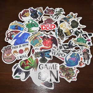 May include: A collection of colourful stickers featuring fantasy and role-playing game themes. The stickers include images of dragons, dice, monsters, and other fantasy creatures. The text on the stickers includes "Ranger", "D&D", "This is how I roll", "House Weapo", and "Game On".