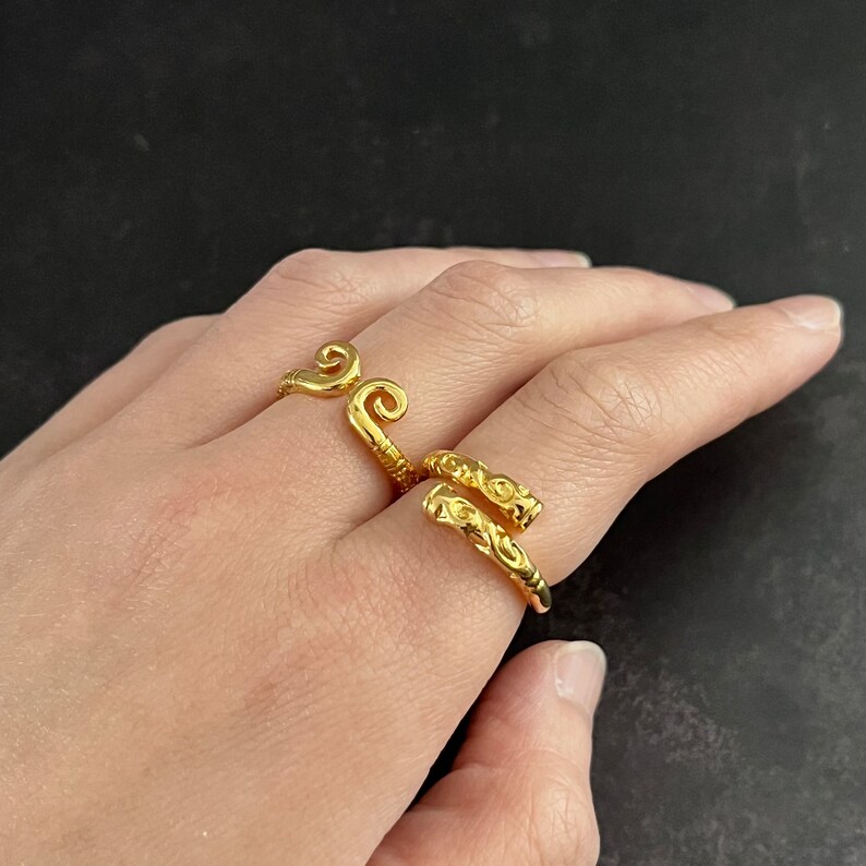 May include: Two gold rings with intricate designs. One ring features a swirling pattern, while the other has a more linear design.