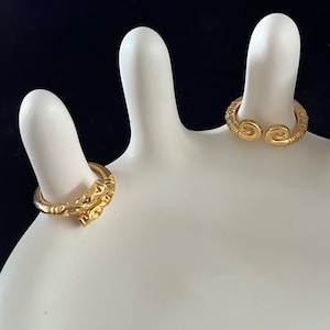 May include: Two gold rings on a white display stand. The ring on the left has a detailed design and the ring on the right is a simple band with a curved design.