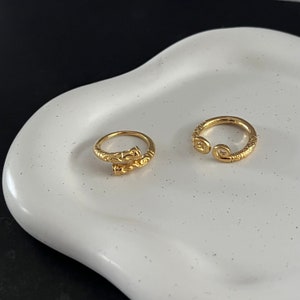 May include: Two gold rings, one with a twisted design and the other with a simple open circle design. The rings are on a white ceramic plate.