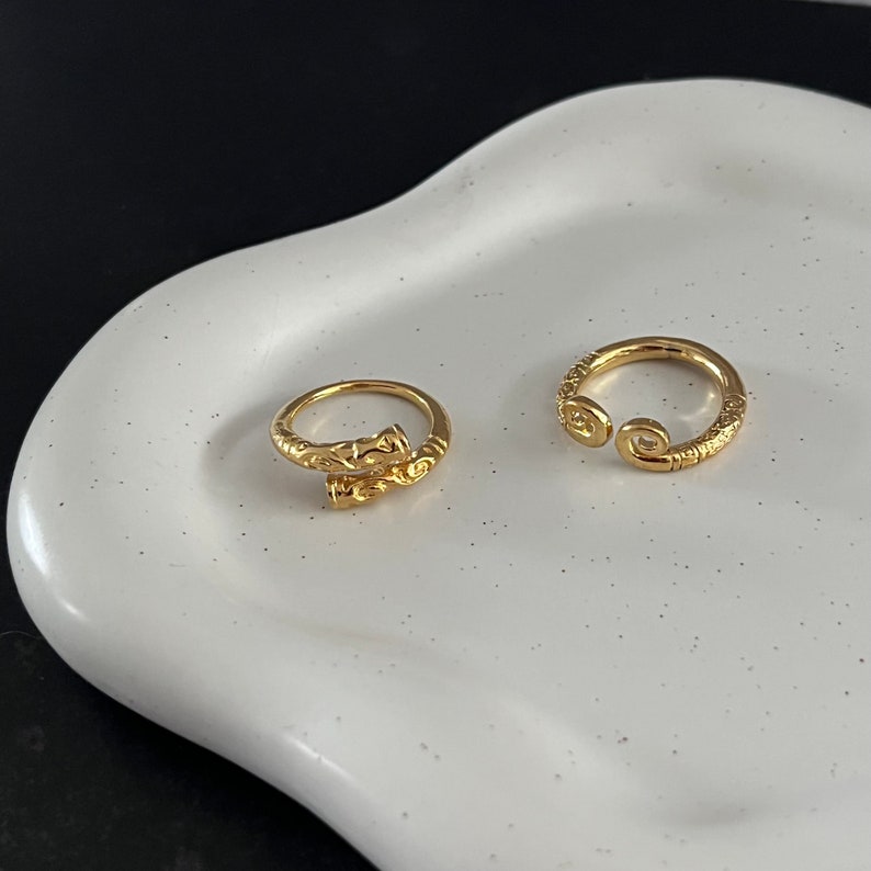 May include: Two gold rings, one with a twisted design and the other with a simple open circle design. The rings are on a white ceramic plate.