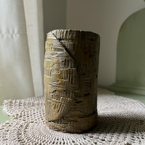 May include: A brown ceramic vase with a textured surface. The vase has a cylindrical shape and is decorated with a woven pattern.