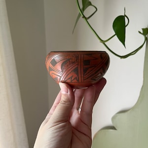 May include: A small, hand-painted ceramic bowl with a geometric design in black and brown on a red background.