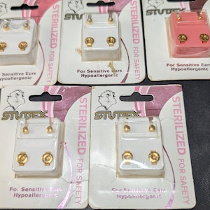 May include: Several packages of Studex hypoallergenic earrings. The earrings are in white and pink plastic packaging with gold-colored accents. The packaging includes the text "STERILIZED FOR SAFETY" and "For Sensitive Ears".