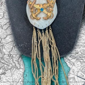 Peut inclure: Collier doré avec un grand pendentif ovale. Le pendentif présente deux ancres croisées, une gemme bleue et une frange de chaîne. Le collier est présenté sur un buste noir sur fond de carte.