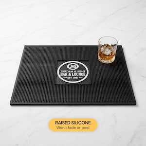 May include: A black bar mat with a textured surface and the text "J&S JORDAN & SONS BAR & LOUNGE EST 1987-". A glass of whiskey with ice sits on the mat. The mat is made of raised silicone. The text "Won't fade or peel" is also visible.