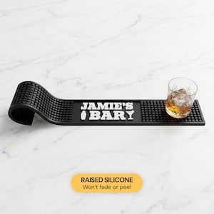 May include: Black silicone bar mat with the text "JAMIE'S BARY" in white. A rocks glass with ice and amber liquid sits on the mat. The mat has a raised, textured surface and a curved end.