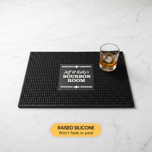 Puede incluir: Alfombrilla de bar negra con superficie texturizada y el texto "Jeff & Kelly's Bourbon Room". Un vaso de whisky con hielo y líquido ámbar está sobre la alfombrilla. La alfombrilla es de silicona en relieve.