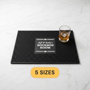 May include: A rectangular black rubber bar mat with a textured nub surface and a central white graphic featuring the text Jeff & Kelly's BOURBON ROOM. A clear glass containing an amber liquid and a single ice cube sits on the right side of the mat.