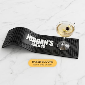 Puede incluir: Alfombrilla de bar negra con el texto "JORDAN'S BAR & CO." en blanco. Un cóctel con una guarnición de cereza está sobre la alfombrilla. La alfombrilla tiene una superficie texturizada y las palabras "RAISED SILICONE" y "Won't fade or peel" están impresas en un óvalo amarillo.