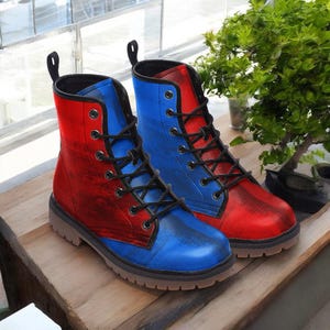 Puede incluir: Un par de botas de combate bicolor. Una bota es roja y azul, la otra es azul y roja. Las botas tienen cordones y ojales negros, con una lengüeta en la parte superior. Las botas están sobre una superficie de madera.