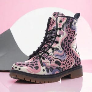 May include: A combat boot with a unique pattern. The boot features a mix of pink, purple, and black leopard print and abstract designs. It has black laces, a black sole, and a brown tread. The boot is a stylish fashion item.