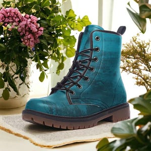 May include: Teal combat boot with black laces and a brown sole. The boot has a textured pattern and a round toe. The boot is displayed on a neutral surface with flowers in the background.