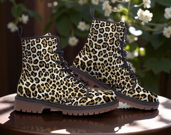 Pode incluir: Um par de botas de tornozelo com estampa de leopardo. As botas têm atacadores pretos, acabamentos pretos e solas castanhas. São exibidas numa mesa castanha com um fundo desfocado de flores brancas e folhagem verde.