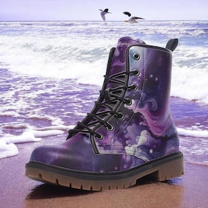 May include: A purple and white galaxy-themed combat boot with black laces and a brown sole. The boot is on a sandy beach with ocean waves in the background. The boot has a unique, artistic design.