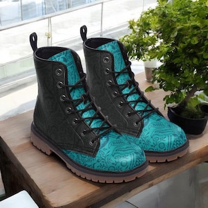 May include: A pair of black and teal combat boots with a floral pattern. The boots have black laces, a pull tab, and a brown sole. The teal section of the boot features a detailed floral design. The boots are displayed on a wooden surface.
