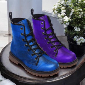 Könnte beinhalten: Ein Paar farbige Kampfstiefel, einer blau und einer lila, mit schwarzen Schnürsenkeln und braunen Sohlen. Die Stiefel sind aus glänzendem Material und werden auf einer dunklen, runden Oberfläche präsentiert. Ein kleiner Blumentopf mit weißen Blüten befindet sich im Hintergrund.