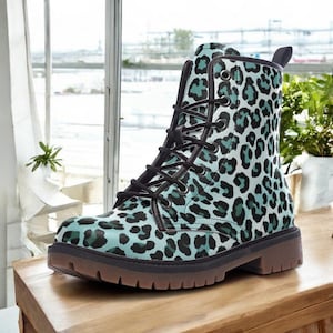 May include: A combat boot with a teal and black leopard print pattern. The boot has black laces, a brown sole, and a pull tab at the back. The boot is sitting on a wooden surface.