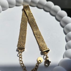 Puede incluir: Una pulsera dorada con un dise&ntilde;o de malla. La pulsera tiene un cierre de cadena y cierre. La pulsera se muestra sobre una superficie blanca con un borde blanco decorativo.