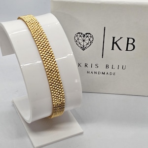 Puede incluir: Una pulsera dorada con un dise&ntilde;o tejido se exhibe en un soporte blanco. La pulsera es rectangular y tiene una superficie texturizada. Una caja blanca con un logotipo de coraz&oacute;n y el texto "KRIS BLIU HANDMADE" est&aacute; en el fondo.