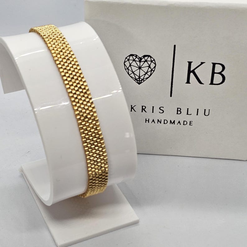 Puede incluir: Una pulsera dorada con un dise&ntilde;o tejido se exhibe en un soporte blanco. La pulsera es rectangular y tiene una superficie texturizada. Una caja blanca con un logotipo de coraz&oacute;n y el texto "KRIS BLIU HANDMADE" est&aacute; en el fondo.