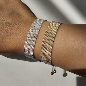 Può includere: Due braccialetti a rete, uno argentato e uno dorato, sono indossati su un polso. I braccialetti hanno un aspetto intrecciato e una consistenza liscia. Il braccialetto dorato ha una delicata catena con due piccole sfere all'estremità per la regolazione.