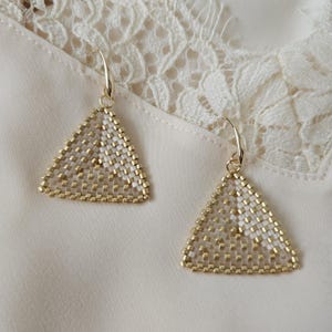 May include: Two gold-coloured, triangular earrings. The earrings are made of small, square beads in gold and white, arranged in a geometric pattern. They hang from gold-coloured ear wires. The background is a cream-coloured fabric.