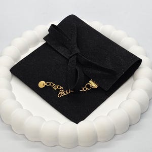 Puede incluir: Un collar de cadena dorada con un peque&ntilde;o colgante y cierre, apoyado sobre una bolsa de terciopelo negro atada con un lazo. La bolsa est&aacute; sobre una bandeja blanca, texturizada y circular.