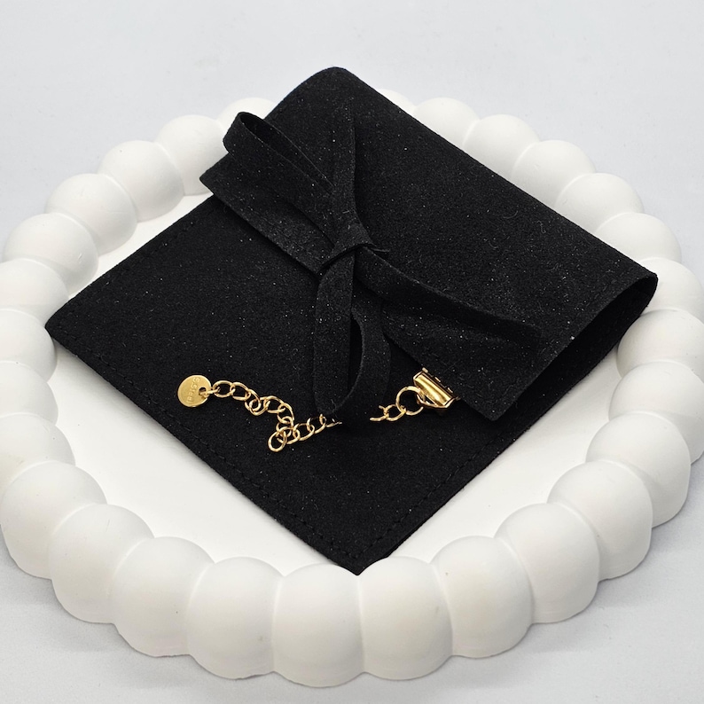 Puede incluir: Un collar de cadena dorada con un peque&ntilde;o colgante y cierre, apoyado sobre una bolsa de terciopelo negro atada con un lazo. La bolsa est&aacute; sobre una bandeja blanca, texturizada y circular.