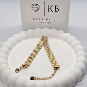 Puede incluir: Una pulsera dorada con un dise&ntilde;o tejido, colocada sobre una superficie blanca texturizada. La pulsera tiene un cierre y una cadena ajustable. El fondo presenta una tarjeta blanca con el texto "KRIS BLIU HANDMADE" y un logotipo de coraz&oacute;n.