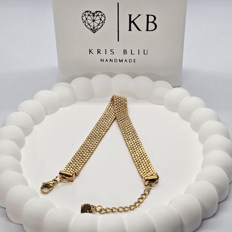 Puede incluir: Una pulsera dorada con un dise&ntilde;o tejido, colocada sobre una superficie blanca texturizada. La pulsera tiene un cierre y una cadena ajustable. El fondo presenta una tarjeta blanca con el texto "KRIS BLIU HANDMADE" y un logotipo de coraz&oacute;n.