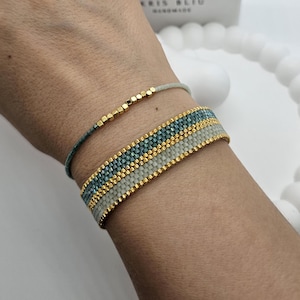 Puede incluir: Dos pulseras de cuentas en una muñeca. La pulsera superior es un hilo delgado de color gris claro con cuentas doradas. La pulsera inferior es una pulsera tejida más ancha con bandas alternas de cuentas grises claras, doradas y azul verdoso.