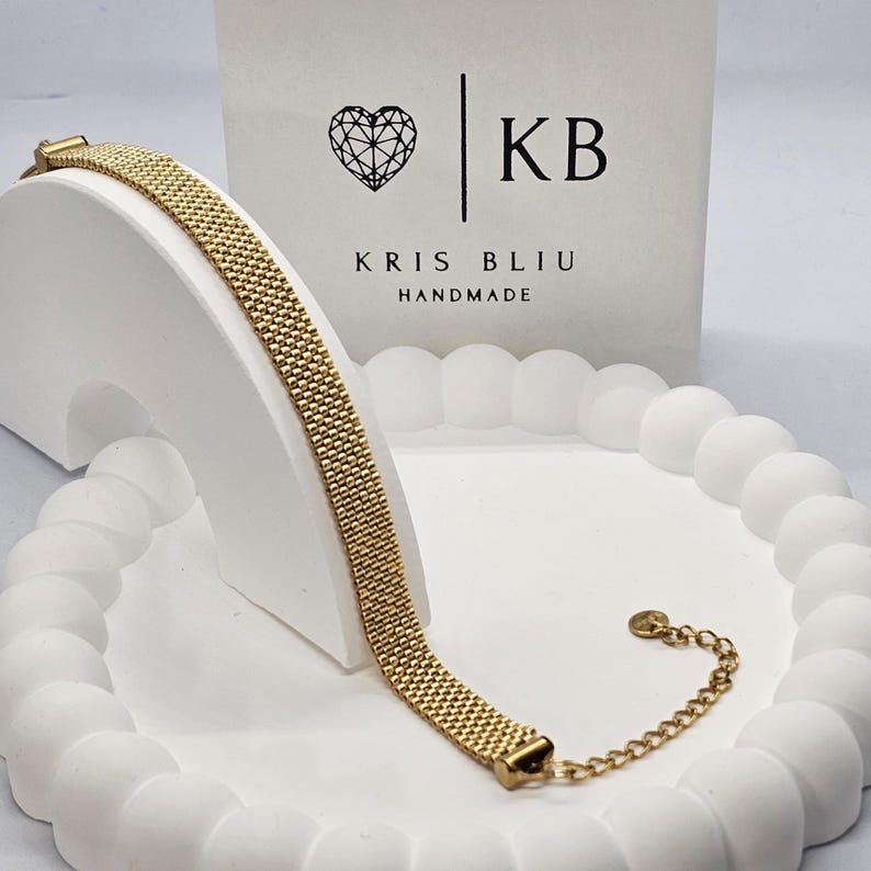 Puede incluir: Una pulsera de malla dorada, exhibida sobre un soporte decorativo blanco. La pulsera tiene forma rectangular con una extensi&oacute;n de cadena. El fondo presenta una caja blanca con un logotipo de coraz&oacute;n y el texto "KB KRIS BLIU HANDMADE."