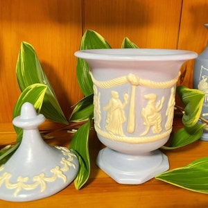 May include: A set of three blue and white ceramic decorative containers with a Greek-inspired design. The containers feature raised white figures and a rope design around the middle. The largest container has a lid with a white ribbon design.