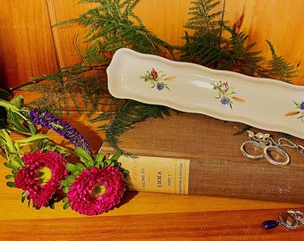 May include: A rectangular, off-white ceramic serving dish with floral designs, resting on a vintage book. The image also features jewelry, including earrings and a bracelet, and fresh flowers. The background is a wooden surface.