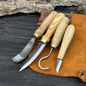 May include: A set of four wood carving tools with wooden handles and metal blades. The tools are arranged on a brown leather surface, with a dark wooden background. The blades have different shapes, including a curved blade and a hook.