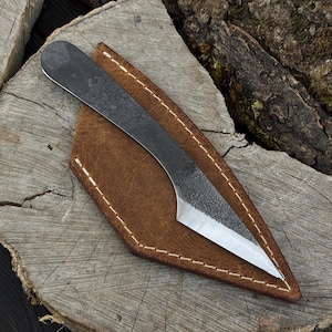 Puede incluir: Un cuchillo de lanzamiento forjado a mano con una hoja de metal oscuro texturizado y una punta afilada. Descansa en una funda de cuero marrón con costuras blancas, sobre un fondo de madera rústica. El cuchillo está diseñado para lanzar y actividades al aire libre.