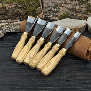 May include: A collection of six wood carving gouges, each with a wooden handle and a metal blade. The tools are displayed in a row, with a leather tool roll in the background. The handles are light-coloured wood, and the blades are silver.